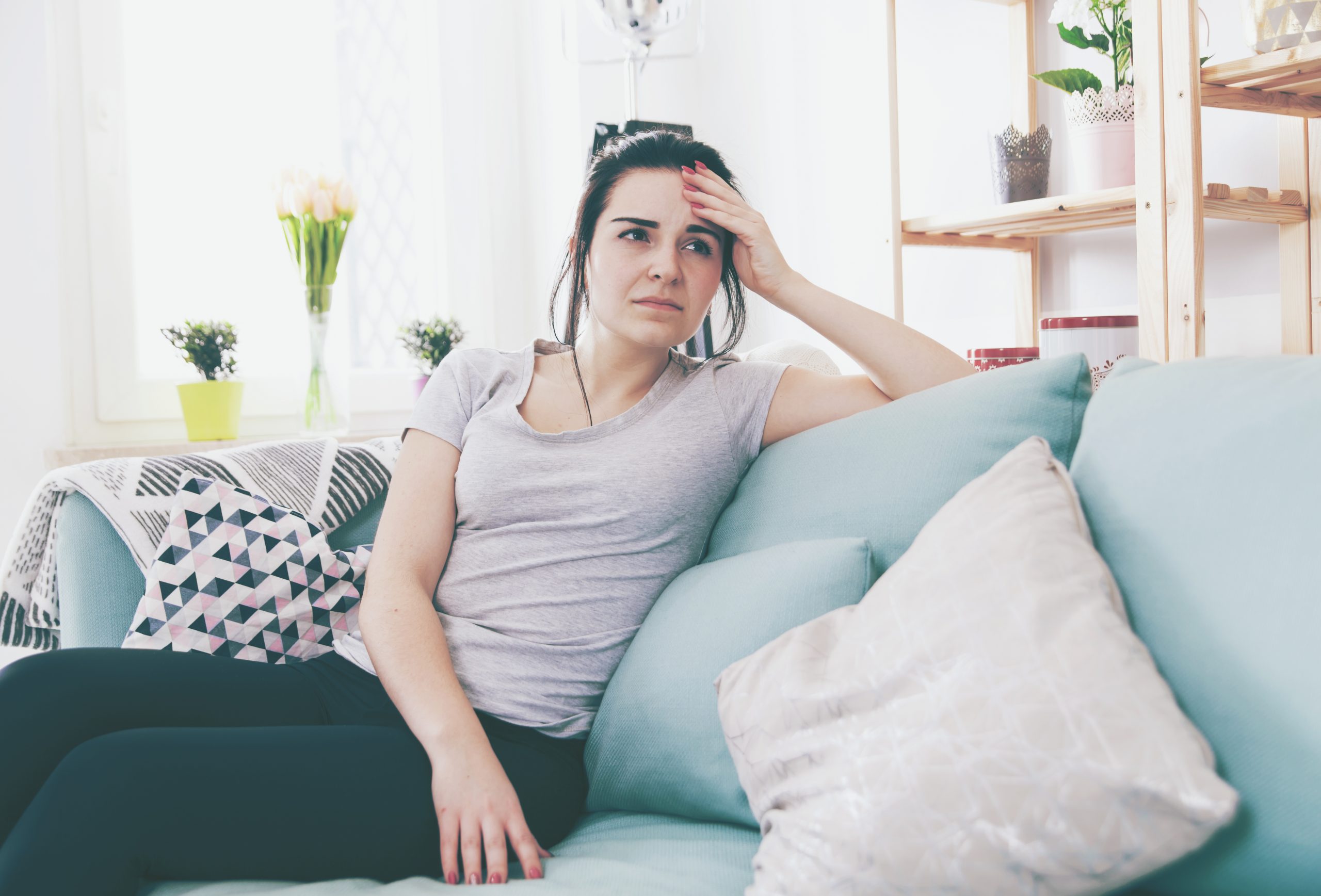 Young worried woman with headache while sitting on comfortable sofa, home interior