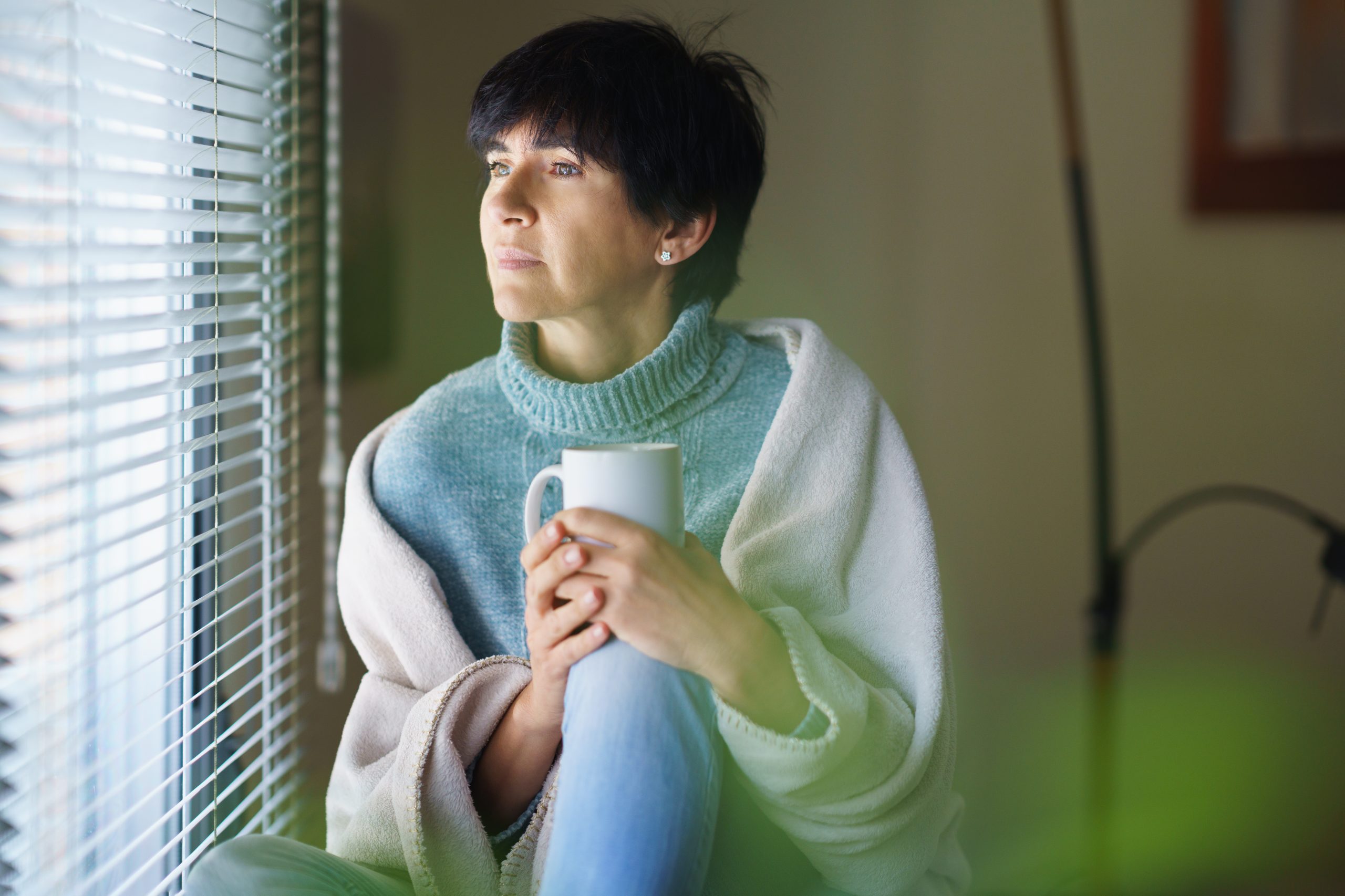 Mid adult woman drinking coffee and looking out of the window in winter day. Female in her 50s