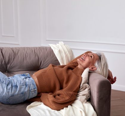 From above of delighted mature female with gray hair and in trendy outfit lying on sofa with hand behind head and laughing while having fun at home